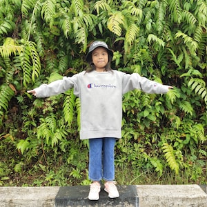 Puede incluir: Una persona joven lleva una sudadera gris con la palabra "Champion" impresa en la parte delantera. Lleva una gorra de cubo negra y pantalones vaqueros azules. La persona está de pie frente a un fondo verde frondoso.