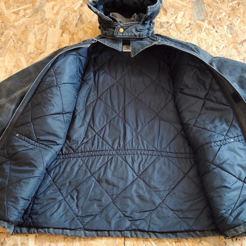 K&ouml;nnte beinhalten: Dunkelgraue Arbeitsjacke mit gestepptem, marineblauem Innenfutter. Die Jacke hat eine Kapuze, einen Rei&szlig;verschluss und einen gekn&ouml;pften Kragen. Das Innere weist eine rautenf&ouml;rmige Steppung auf.