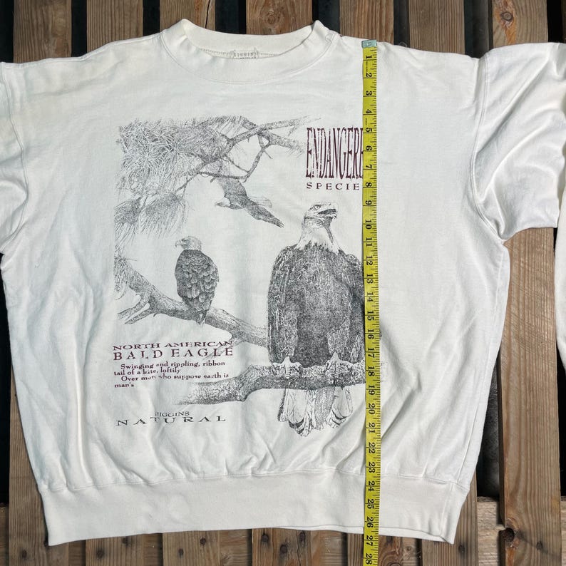 May include: White long-sleeve sweatshirt with a graphic of a bald eagle on a branch. The text "ENDANGERED SPECIES" and "NORTH AMERICAN BALD EAGLE" is printed on the shirt. The text "MOGINS NATURAL" is printed at the bottom of the shirt.