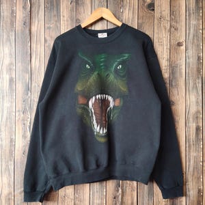 Op de afbeelding: Zwart sweatshirt met een grote afbeelding van een dinosaurusgezicht in groen- en bruintinten. De mond van de dinosaurus is open en toont witte tanden en een rood interieur. Het sweatshirt hangt aan een houten hanger.