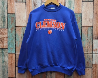 May include: A royal blue sweatshirt with the word "CLEMSON" in orange and white lettering across the chest. Below the word "CLEMSON" is the text "AUTHENTIC SPORT" in white lettering. A paw print is below the text. The sweatshirt is hanging on a wooden hanger.