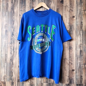 May include: A blue t-shirt with a green and white Seattle Seahawks logo. The logo says "Seattle" in a large green and white font, and "National Football League" in a smaller green font. The logo also includes a green and white wreath with the words "Seahawks Member Club NFL" inside.