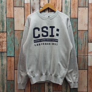 May include: A light gray crewneck sweatshirt featuring the dark navy blue text CSI: CRIME SCENE INVESTIGATION LAS VEGAS UNIT printed on the chest. The garment has long sleeves, a ribbed collar, cuffs, and hem, and a small triangular stitch detail.