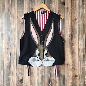 Puede incluir: Chaleco negro con diseño de Bugs Bunny. El chaleco tiene cuello en V, cierre de botones y forro a rayas. La cara de Bugs Bunny se muestra prominentemente en la parte delantera.