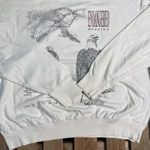 May include: White long-sleeve sweatshirt with a black and white illustration of an eagle. The text "ENDANGERED SPECIES" is printed in maroon on the front of the shirt. The text "MIGRINS NATURA" is printed in black on the bottom of the shirt.