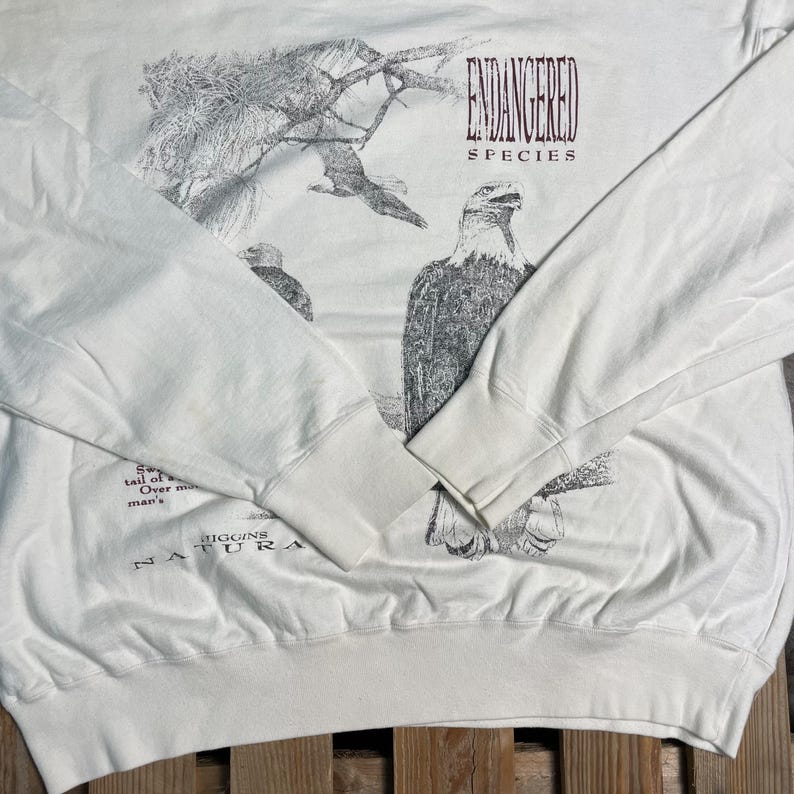May include: White long-sleeve sweatshirt with a black and white illustration of an eagle. The text "ENDANGERED SPECIES" is printed in maroon on the front of the shirt. The text "MIGRINS NATURA" is printed in black on the bottom of the shirt.