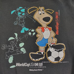 May include: A black t-shirt with a gold embroidered image of a cartoon dog wearing a red and blue soccer uniform and kicking a soccer ball. The dog is surrounded by other cartoon dogs playing soccer. The text "World Cup USA94" is embroidered in gold on the shirt.