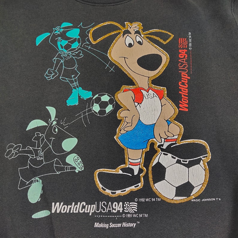 May include: A black t-shirt with a gold embroidered image of a cartoon dog wearing a red and blue soccer uniform and kicking a soccer ball. The dog is surrounded by other cartoon dogs playing soccer. The text "World Cup USA94" is embroidered in gold on the shirt.