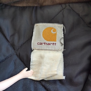 K&ouml;nnte beinhalten: Nahaufnahme eines Carhartt-Etiketts mit einem goldenen Halbmond und dem Markennamen in roter Schrift. Das Etikett ist an einem schwarzen, gesteppten Stoff befestigt, mit einem braunen Cordkragen oben.