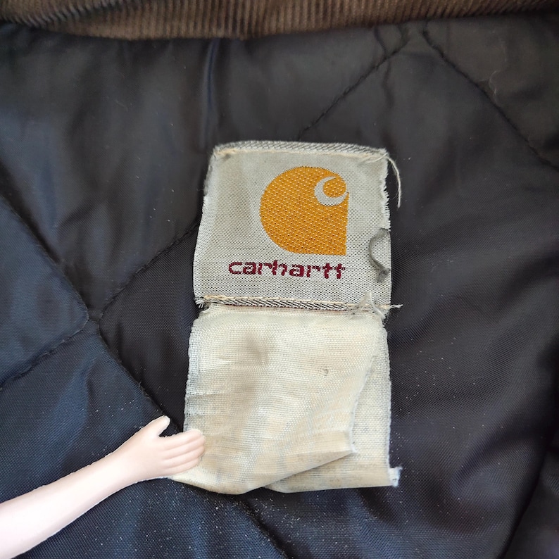 K&ouml;nnte beinhalten: Nahaufnahme eines Carhartt-Etiketts mit einem goldenen Halbmond und dem Markennamen in roter Schrift. Das Etikett ist an einem schwarzen, gesteppten Stoff befestigt, mit einem braunen Cordkragen oben.