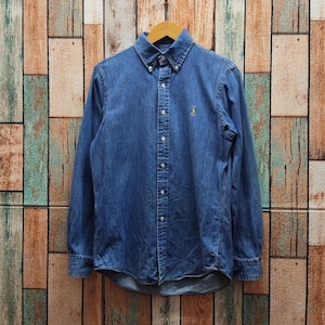 May include: A long-sleeved denim shirt in a medium blue wash, buttoned up and hanging on a wooden hanger. The shirt has a button-down collar and a small embroidered logo on the chest. The background is a wooden wall with vertical planks.