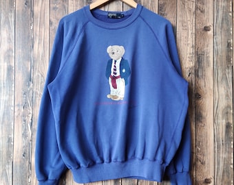 Puede incluir: Una sudadera azul con un logotipo de Polo Ralph Lauren y un oso que lleva una chaqueta azul, corbata roja y pantalones blancos. El oso sostiene una raqueta de tenis.