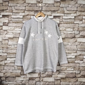 May include: Gray sweatshirt with long sleeves and a white collar. The sweatshirt has a white stripe on each sleeve and a white embroidered design on the front that says "Sport Club College" with five white stars.