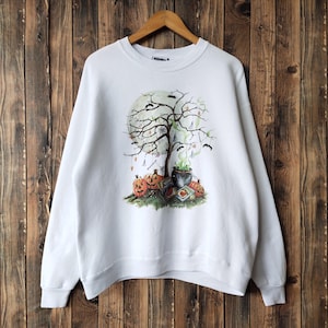 May include: White Halloween-themed sweatshirt featuring a spooky tree, pumpkins, bats, and a cauldron with green smoke. The design is centered on the front of the long-sleeved crew neck shirt. The sweatshirt is hanging on a wooden hanger.