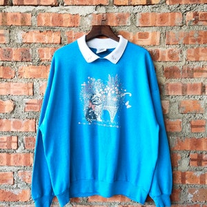 May include: A blue sweatshirt with a white collar and a print of a cat in a wheelbarrow with flowers.
