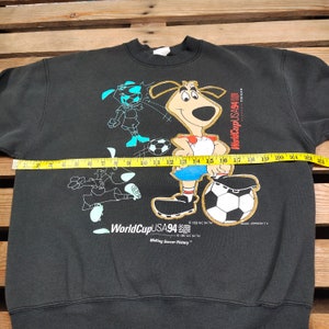 May include: Black sweatshirt with a graphic print of a cartoon dog and a soccer ball. The text "World Cup USA 94" and "Making Soccer History" is printed on the sweatshirt.