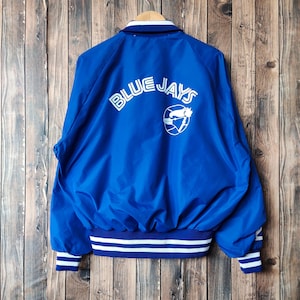 May include: A blue Blue Jays jacket with white lettering and a logo on the back. The jacket has a white and blue striped collar, cuffs, and waistband. The jacket is hanging on a wooden hanger.