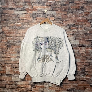 Pode incluir: Uma camisa de moletom cinza com uma estampa gráfica de uma casa com uma varanda e degraus. A casa é cercada por árvores e há um caminho que leva até a varanda.