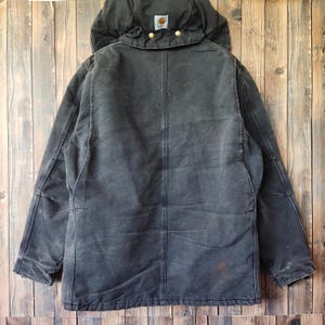K&ouml;nnte beinhalten: Schwarze Kapuzenjacke mit abgenutztem Aussehen. Die Jacke hat eine gro&szlig;e Kapuze, messingfarbene Kn&ouml;pfe und einen kleinen quadratischen Logoaufn&auml;her. Die Jacke ist aus strapazierf&auml;higem Material gefertigt und hat eine Arbeitskleidungs&auml;sthetik.
