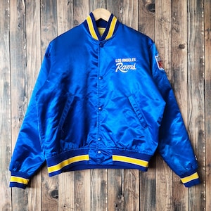 Puede incluir: Una chaqueta bomber de satén azul con ribetes de rayas amarillas y blancas. La chaqueta tiene cierre de presión y presenta el logotipo de los Los Angeles Rams en el pecho izquierdo.