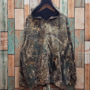 May include: A camouflage-patterned zip-up hoodie with a hood. The jacket features a brown, green, and tan woodland camouflage design. It has a front pocket and a Carhartt logo patch. The hoodie is hanging on a wooden hanger.