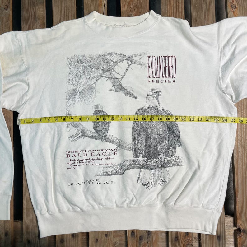 May include: White long-sleeve sweatshirt with a graphic print of a bald eagle and the text "ENDANGERED SPECIES NORTH AMERICAN BALD EAGLE" and "BIG GAME NATURAL".