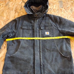K&ouml;nnte beinhalten: Dunkelgraue Carhartt Kapuzenjacke mit gelbem Ma&szlig;band auf der Vorderseite. Die Jacke hat zwei Brusttaschen, zwei untere Taschen und einen Carhartt-Logoaufn&auml;her. Die Jacke misst etwa 69 cm quer.