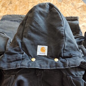 K&ouml;nnte beinhalten: Nahaufnahme einer dunkelgrauen Carhartt Kapuzenjacke. Die Kapuze ist hochgeklappt und zeigt das Carhartt-Logo-Patch in der Mitte. Zwei goldfarbene Kn&ouml;pfe sind unterhalb der Kapuze sichtbar. Die Jacke scheint aus strapazierf&auml;higem Workwear-Stoff zu sein.