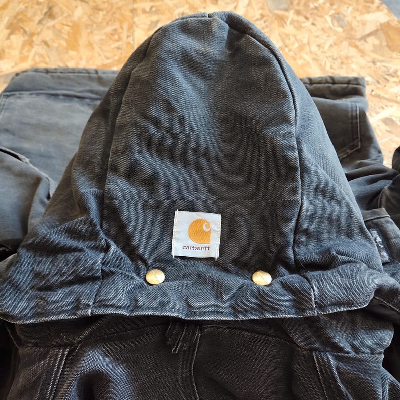 K&ouml;nnte beinhalten: Nahaufnahme einer dunkelgrauen Carhartt Kapuzenjacke. Die Kapuze ist hochgeklappt und zeigt das Carhartt-Logo-Patch in der Mitte. Zwei goldfarbene Kn&ouml;pfe sind unterhalb der Kapuze sichtbar. Die Jacke scheint aus strapazierf&auml;higem Workwear-Stoff zu sein.
