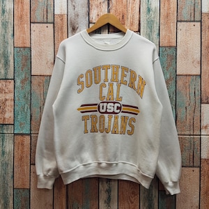 Puede incluir: Una sudadera blanca de cuello redondo con el texto SOUTHERN CAL TROJANS en amarillo con borde granate. El centro incluye un gráfico granate y amarillo con las letras USC dentro de un círculo, flanqueado por líneas horizontales sobre fondo blanco.
