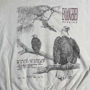 May include: A white t-shirt with a black and white illustration of a bald eagle perched on a branch. The text "ENDANGERED SPECIES" is printed in maroon above the eagle, and "NORTH AMERICAN BALD EAGLE" is printed below the eagle. The text "IGGINS NATURAL" is printed in maroon at the bottom of the shirt.