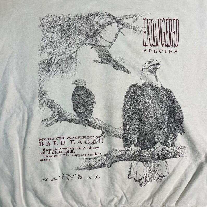 May include: A white t-shirt with a black and white illustration of a bald eagle perched on a branch. The text "ENDANGERED SPECIES" is printed in maroon above the eagle, and "NORTH AMERICAN BALD EAGLE" is printed below the eagle. The text "IGGINS NATURAL" is printed in maroon at the bottom of the shirt.
