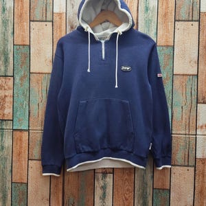 May include: A navy blue hooded sweatshirt with a white-lined hood and hem. The sweatshirt features a front pocket, a quarter-zip closure, and a small patch on the chest. A small red and white tag is on the sleeve.