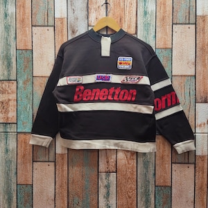 May include: Black long-sleeve sweatshirt with white stripes and the word "Benetton" in red. The shirt has patches with text and logos, including "UCB Power 500 Racing". The shirt is hanging on a wooden hanger.