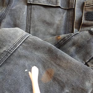 K&ouml;nnte beinhalten: Nahaufnahme einer abgenutzten, dunkelgrauen Jeansjacke. Die Jacke hat sichtbare N&auml;hte, eine Tasche und einen Klettverschluss. Es gibt mehrere Flecken und Markierungen auf dem Stoff, die auf Abnutzung und Gebrauch hindeuten. Das Gesamtbild deutet auf einen Vintage- oder Arbeitskleidungsstil hin.