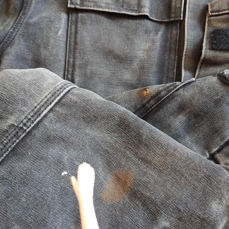 K&ouml;nnte beinhalten: Nahaufnahme einer abgenutzten, dunkelgrauen Jeansjacke. Die Jacke hat sichtbare N&auml;hte, eine Tasche und einen Klettverschluss. Es gibt mehrere Flecken und Markierungen auf dem Stoff, die auf Abnutzung und Gebrauch hindeuten. Das Gesamtbild deutet auf einen Vintage- oder Arbeitskleidungsstil hin.