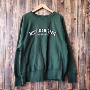 Puede incluir: Sudadera verde oscuro con las palabras "MICHIGAN STATE" en letras blancas arqueadas en el pecho. Debajo está la palabra "SPARTANS". La sudadera tiene cuello redondo y mangas largas. Cuelga de una percha de madera.