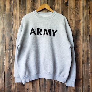 May include: A light gray sweatshirt with the word "ARMY" printed in large, black capital letters across the chest. The sweatshirt is hanging on a wooden hanger against a backdrop of vertical wooden planks.