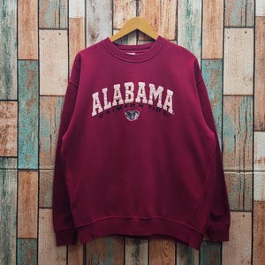 May include: A maroon long-sleeve crewneck sweatshirt featuring the word ALABAMA in large white block letters arched across the chest, with the words CRIMSON TIDE and a small elephant graphic printed in black directly below the main text.
