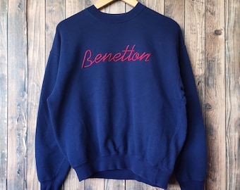 Puede incluir: Una sudadera azul marino con la palabra "Benetton" bordada en rojo en el pecho. La sudadera está colgada de una percha de madera sobre un fondo de tablones de madera. La prenda tiene cuello redondo y mangas largas.