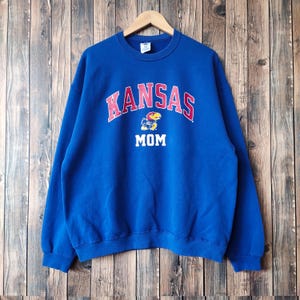 Pode incluir: Camisola azul com a palavra "KANSAS" em letras vermelhas e brancas com um pequeno logotipo de mascote e a palavra "MOM" por baixo.