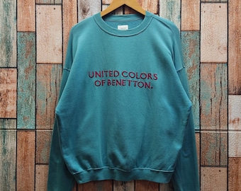 May include: A teal long-sleeve crewneck sweatshirt featuring the text UNITED COLORS OF BENETTON. embroidered in dark red on the chest. The garment has a ribbed collar, cuffs, and hem, and is displayed on a wooden hanger.