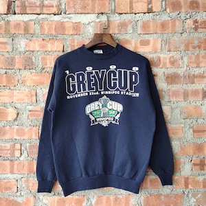 Puede incluir: Una sudadera azul marino con el texto "1998 Grey Cup" y "November 22nd. Winnipeg Stadium" impreso en la parte delantera. La sudadera presenta un gráfico de un trofeo con el texto "Grey Cup 1998" y "Winnipeg" debajo.