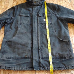 K&ouml;nnte beinhalten: Eine dunkelgraue Arbeitsjacke mit Kapuze, zwei Brusttaschen und zwei gro&szlig;en unteren Taschen. Die Jacke ist etwa 76 cm lang, gemessen mit einem gelben Ma&szlig;band. Das Carhartt-Logo ist auf der Brusttasche sichtbar.