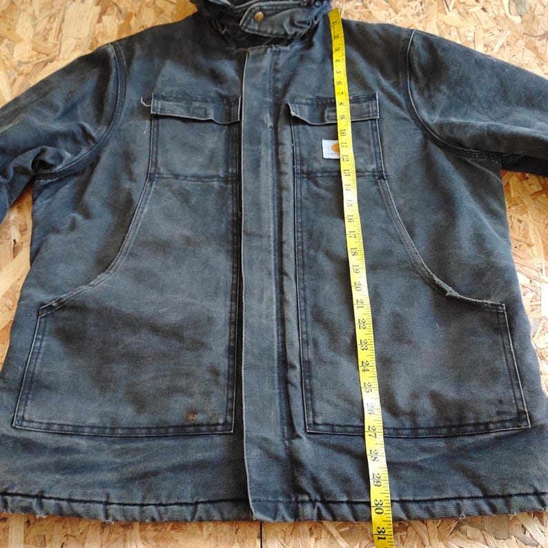 K&ouml;nnte beinhalten: Eine dunkelgraue Arbeitsjacke mit Kapuze, zwei Brusttaschen und zwei gro&szlig;en unteren Taschen. Die Jacke ist etwa 76 cm lang, gemessen mit einem gelben Ma&szlig;band. Das Carhartt-Logo ist auf der Brusttasche sichtbar.