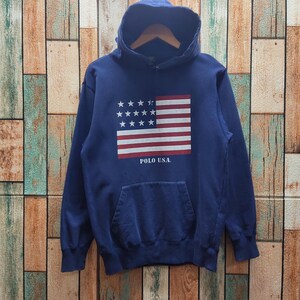 May include: Navy blue hooded sweatshirt with a front pocket and a graphic of the American flag. The flag design features white stars and red and white stripes. The text "POLO U.S.A." is printed below the flag.
