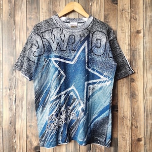 Puede incluir: Una camiseta azul y negra con una gran estrella blanca y las palabras "Dallas Cowboys" impresas en ella. La camiseta tiene un aspecto descolorido y desgastado.