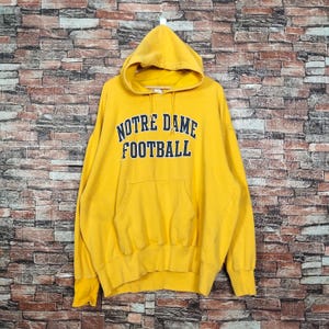Puede incluir: Sudadera amarilla con capucha con el texto "Notre Dame Football" en letras azules.