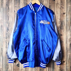 Könnte beinhalten: Eine blaue Satin-Baseballjacke mit weißen und grauen Ärmeln. Die Jacke hat einen Reißverschluss und ein weiß gesticktes Logo mit der Aufschrift "Orlando Magic" und einem Basketball.