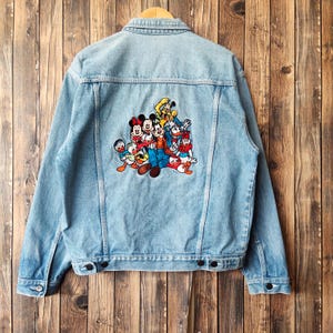 Puede incluir: Chaqueta vaquera azul claro con un diseño bordado colorido de personajes de Disney en la espalda. La chaqueta tiene un diseño clásico con botones, dos bolsillos en el pecho y un cuello. La chaqueta está colgada en una percha de madera.
